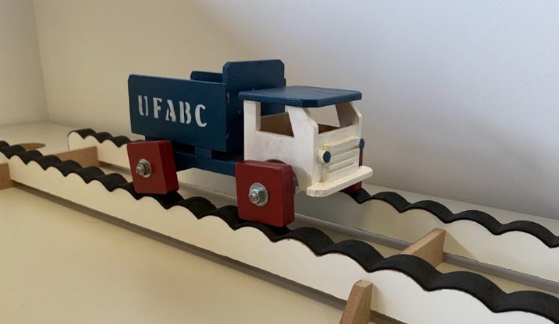 Na imagem temos um mini caminhão branco de madeira, cuja caçamba é azul escura e está escrito em letras brancas “UFABC”. As rodas do caminhão são vermelhas e quadradas. Cada par de rodas do caminhão está em cima de uma pista com ondulações.